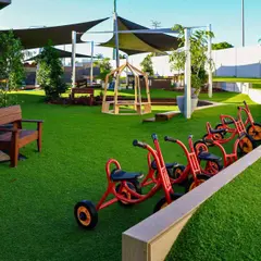 Synthetic grass yard with red trikes, timber bench seats and shade sails at a childcare centre
