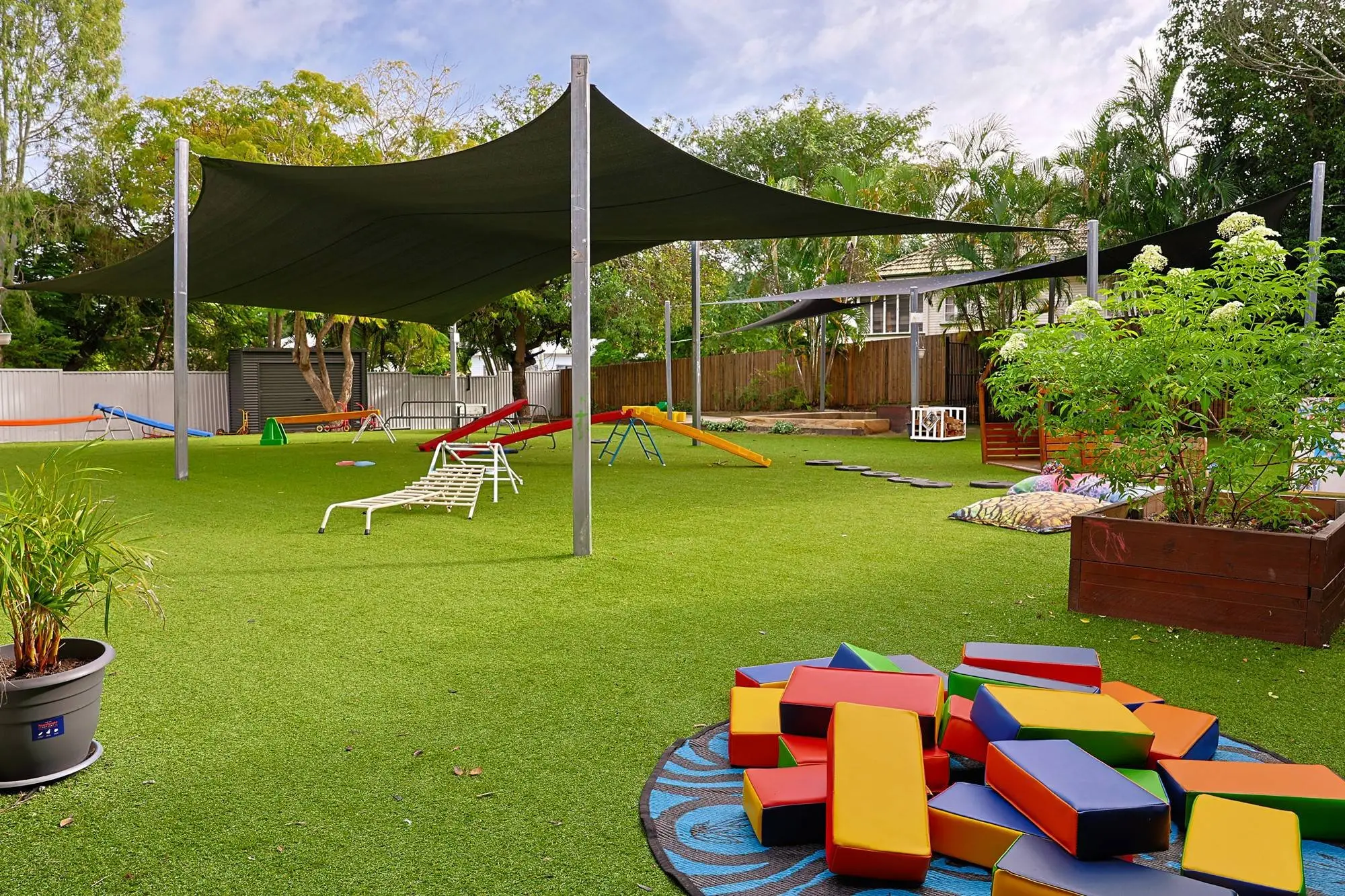 Shade sails over a synthetic grass play yard with soft play blocks at a childcare centre