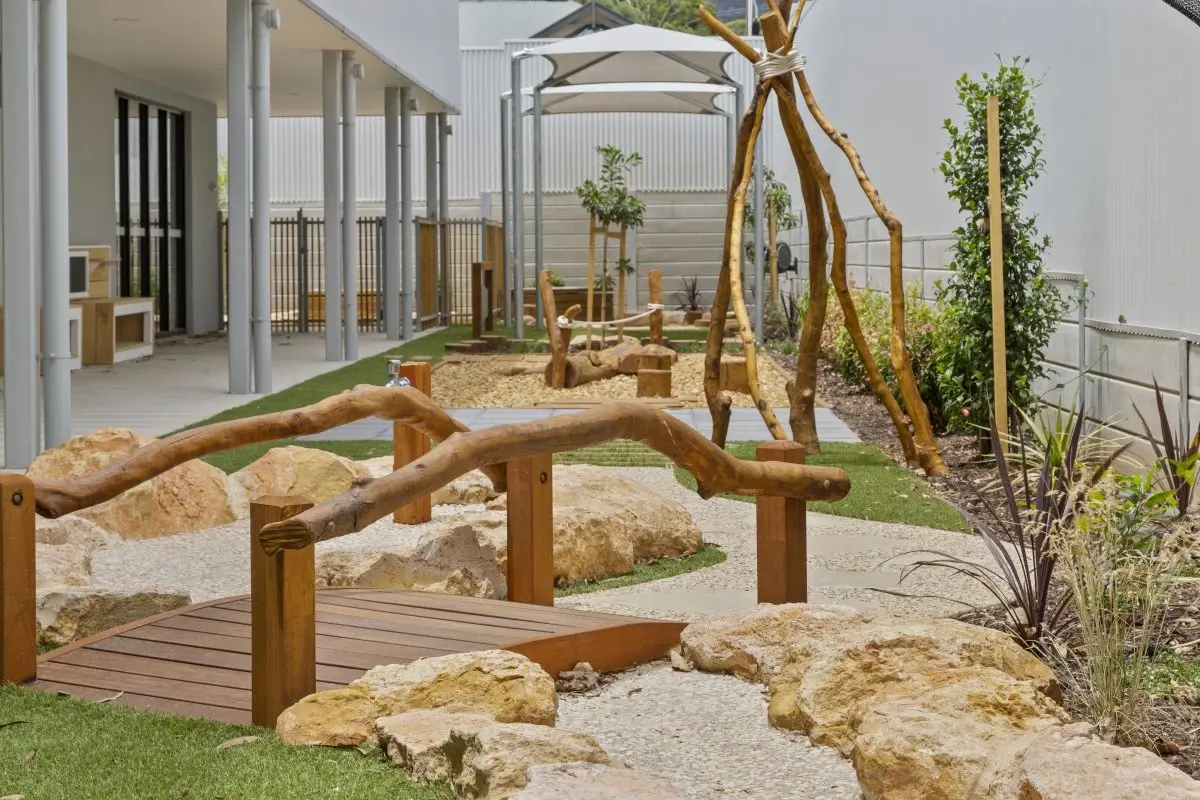 Nature play yard with a timber bridge, sandstone boulders and a cubby at a childcare centre