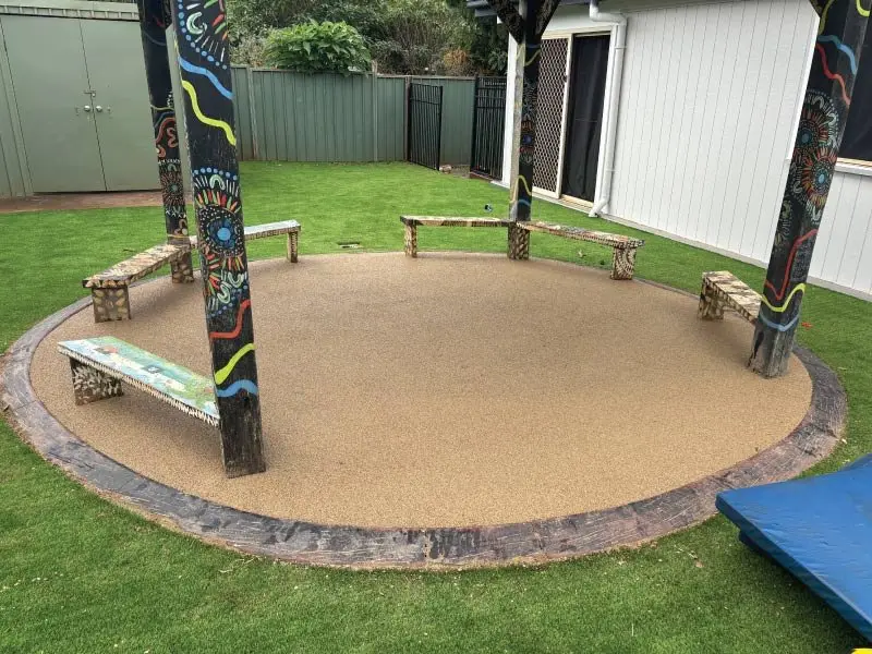 Circular wetpour softfall pad with timber bench seating under a painted shelter at a childcare playground